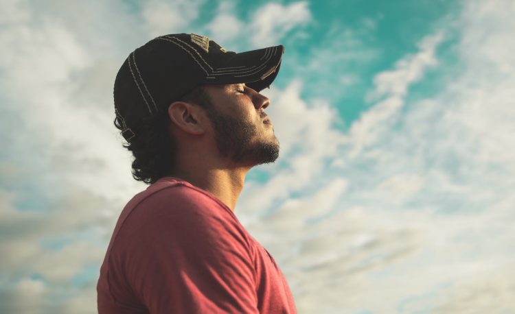 man wearing black cap with eyes closed under cloudy sky